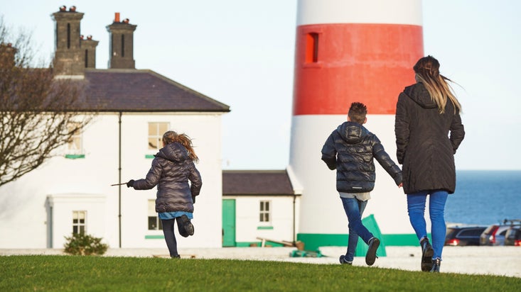 A woman and two children facing away from the camera, walking towards a white house and large white and red lighthouse.